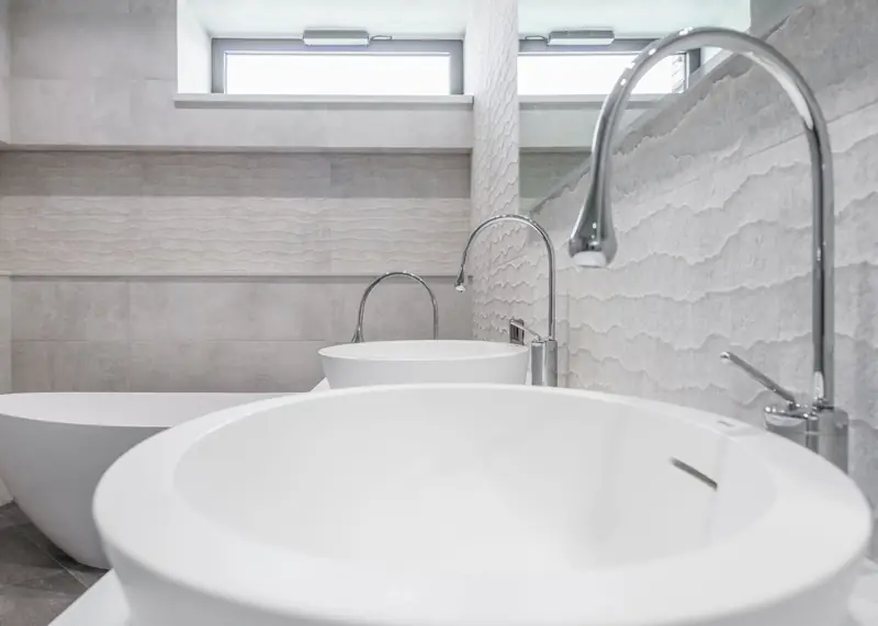 A freestanding tub and a round vessel basin against textured pale stone tiling.