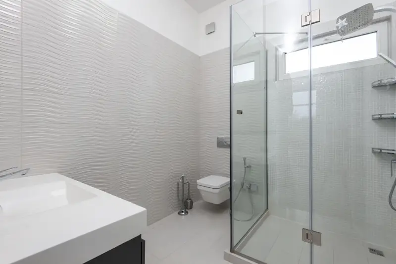 A bathroom in wave-textured pale tile with a glass corner shower and wall-hung toilet.